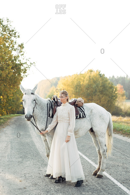 Blonde woman dressed in vintage dress standing on country road with horse