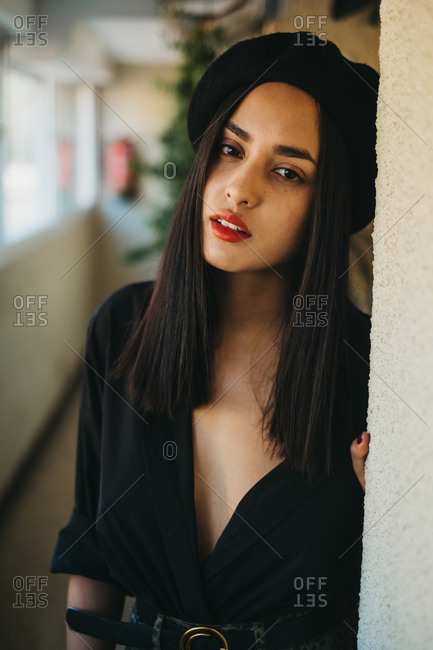Attractive young woman in black beret looking at camera while leaning on wall at home