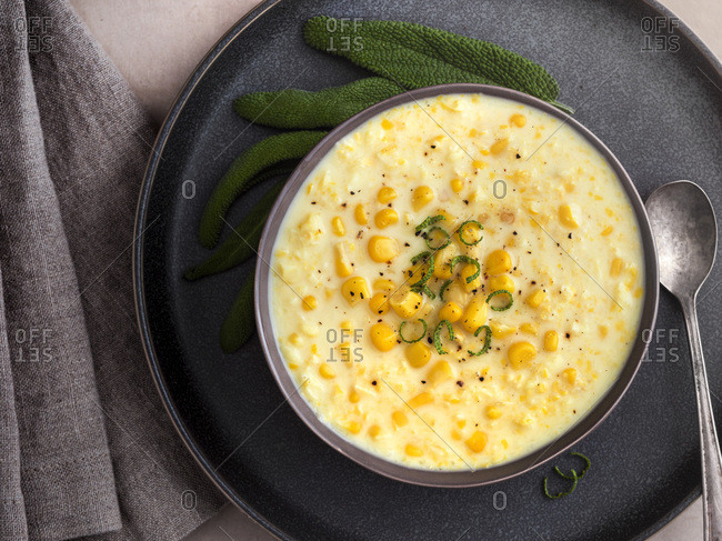 A bowl of warm, sweet, corn soup. Gray bowl and plate garnished with whole and chopped sage on a concrete surface. Warm tones.