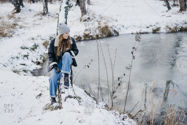 beautiful woman sitting on a tree and looking at the river. on the background of winter forest and river