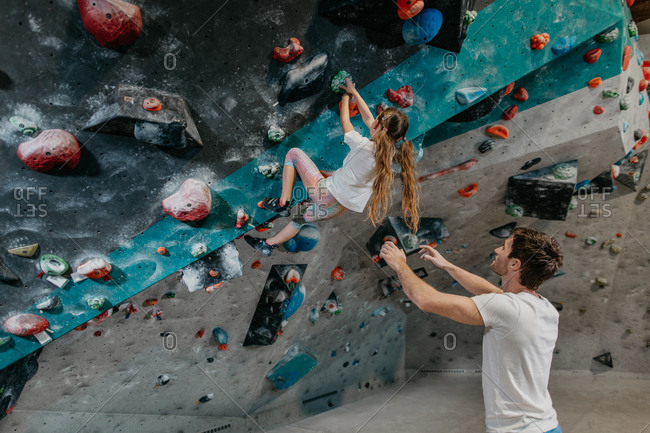 Young girl making her way up an artificial climbing wall with an instructor spotting. Child learning to climb up a bouldering wall with a coach in an indoor bouldering gym.