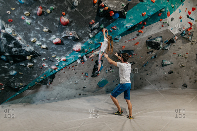 Back view of a bouldering instructor catching a child hanging from a climbing wall. Child learning to climb up a bouldering wall with a coach in an indoor bouldering gym.