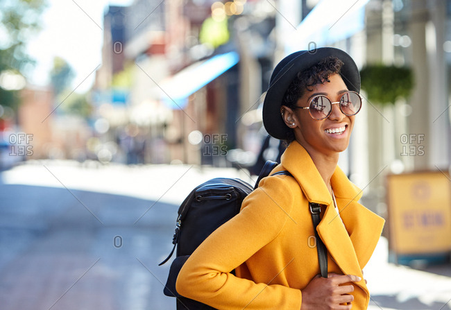 Close up of young woman wearing black homburg hat,  sunglasses, yellow coat and a backpack standing on a city street smiling to camera, waist up, side view