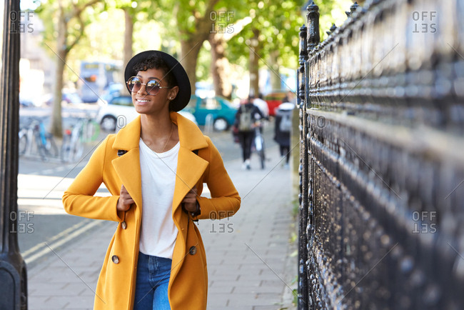 Fashionable young woman wearing black homburg hat,  sunglasses and unbuttoned yellow coat and blue jeans walking on a city street towards camera past railings, close up, selective focus