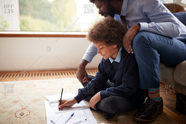 Pre-teen boy in school uniform sitting on the floor at home doing his homework helped by his father, sitting behind him on sofa, side view, close up
