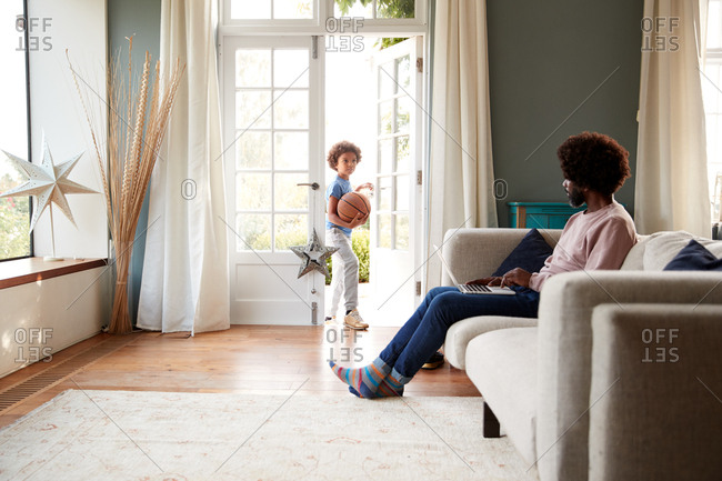Middle aged man sitting on a sofa in living room using a laptop talks with his son, who stands at the open French window holding a basketball, selective focus