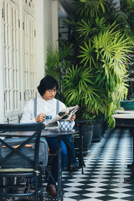 Serious middle-aged Asian businessman having a breakfast at hotel terrace and reading newspaper while drinking a tea.