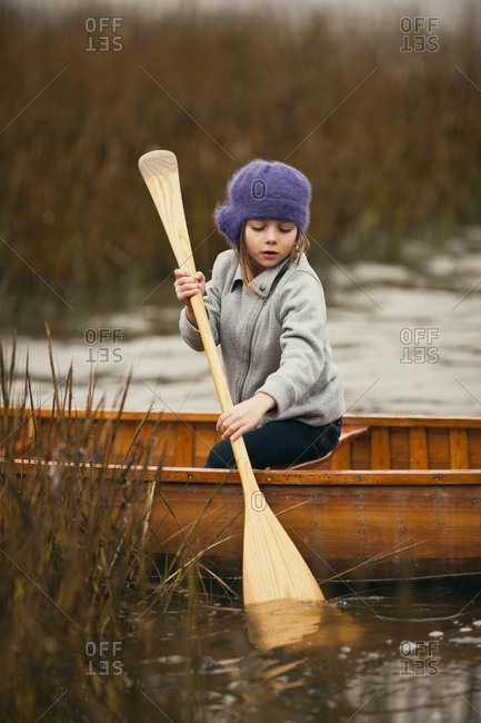 Young girl looks down as she tries to paddle a wooden canoe through reeds in a lake.