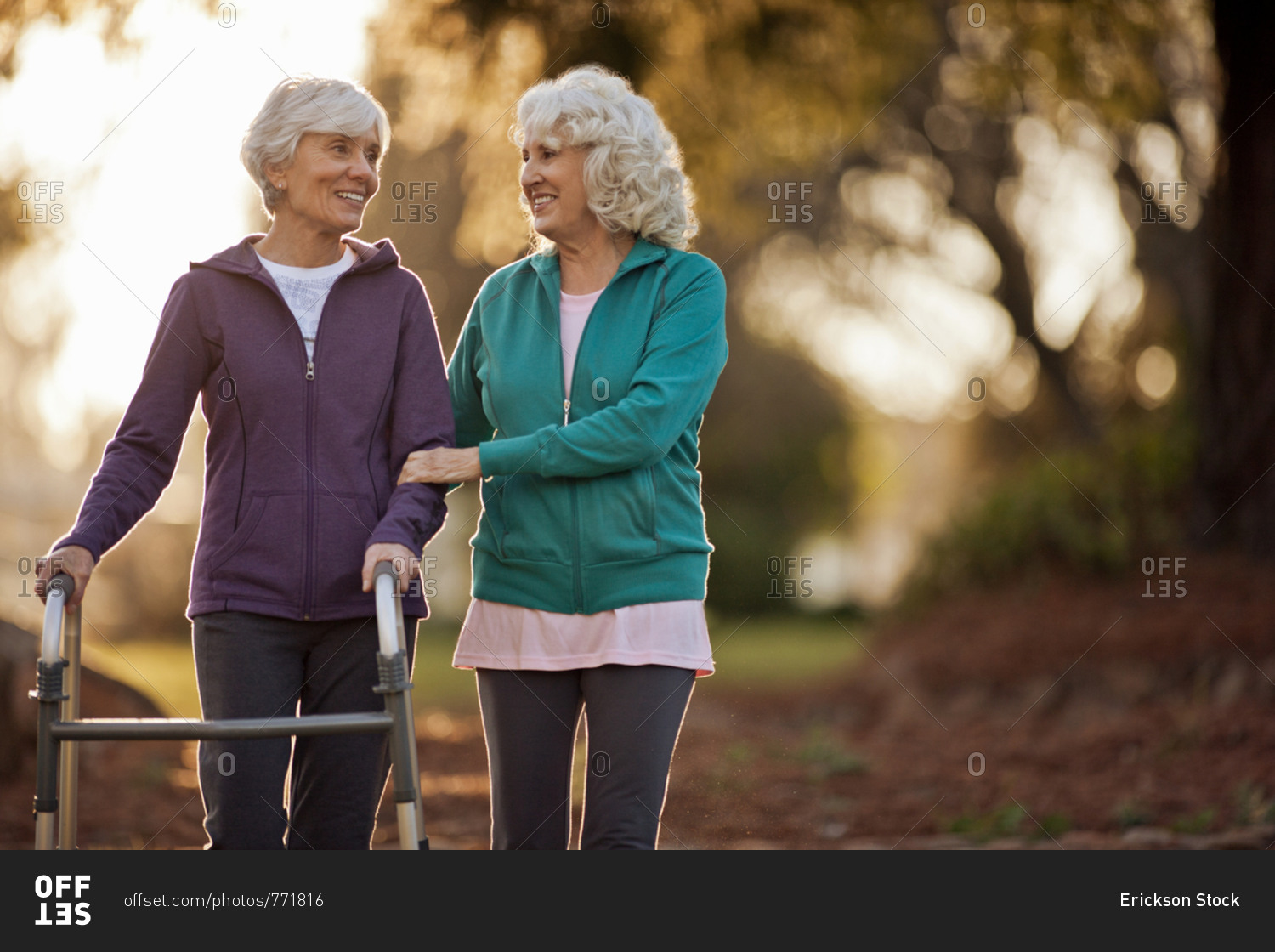 Smiling senior woman helping her friend to walk with a walking aid through a park.