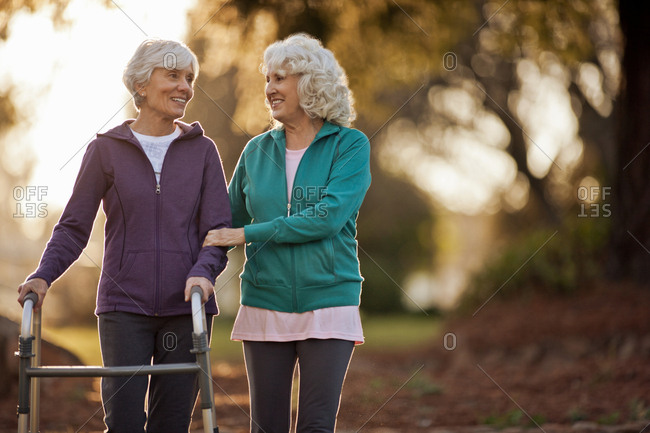 Smiling senior woman helping her friend to walk with a walking aid through a park.