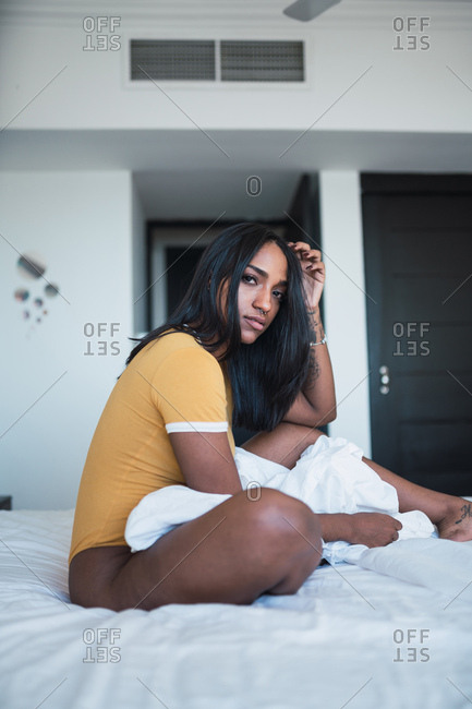 Side view of sensual African American lady looking at camera and scratching head while sitting on soft bed in stylish room