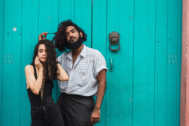 Young Hispanic man with hairs on hand and woman with hands on head in casual wear looking at camera near blue door on street in Colonial Zone in Santo Domingo city, Dominican Republic