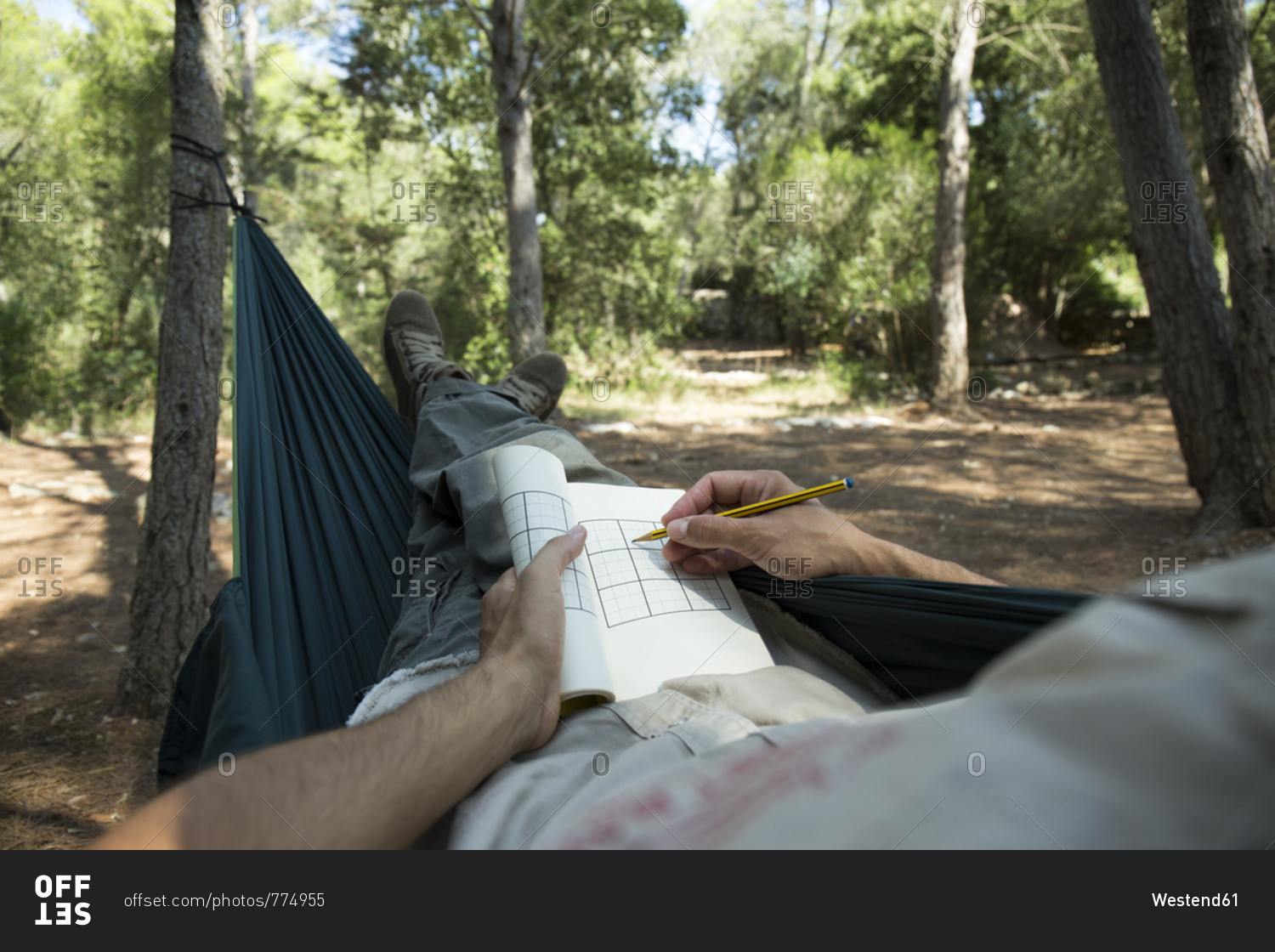 Man lying in hammock in the woods relaxing with puzzle book- partial view