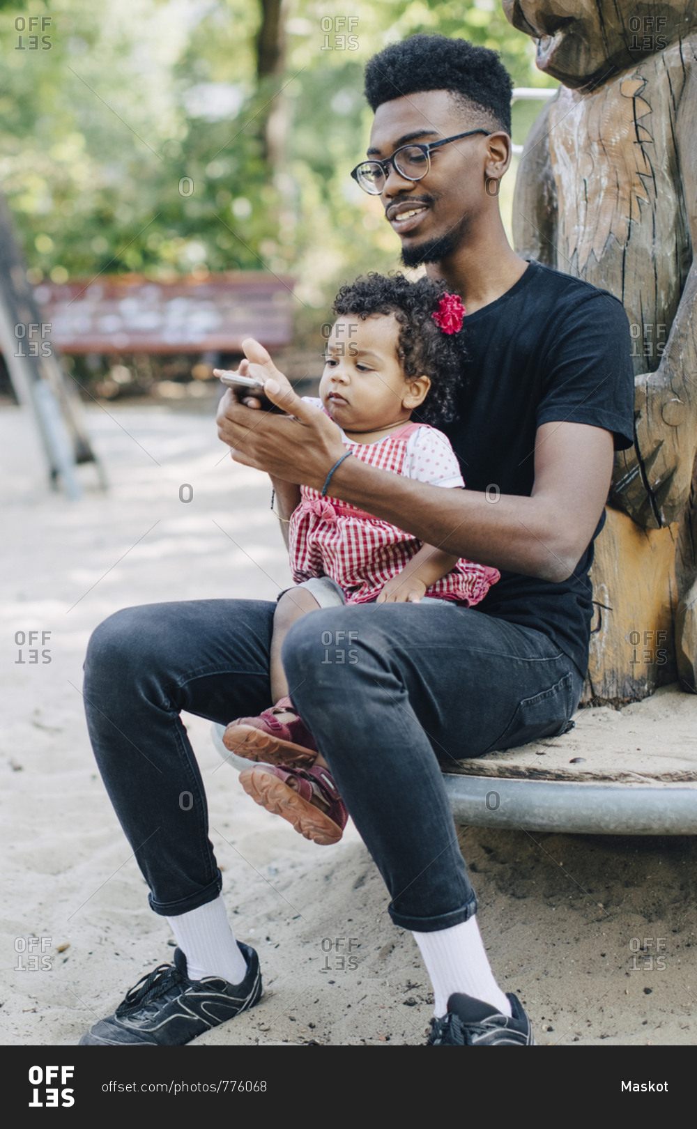 Baby girl using mobile phone while sitting with father on outdoor play equipment at playground