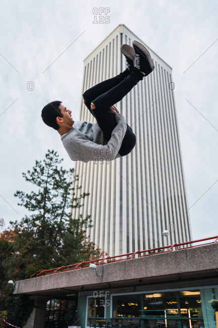 Side view of handsome young guy performing side flip while doing parkour against modern building and cloudy sky on street of Madrid
