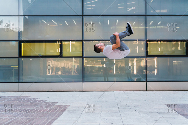Side view of handsome young male in stylish outfit performing back flip while doing parkour near glass wall of modern building on street of Madrid