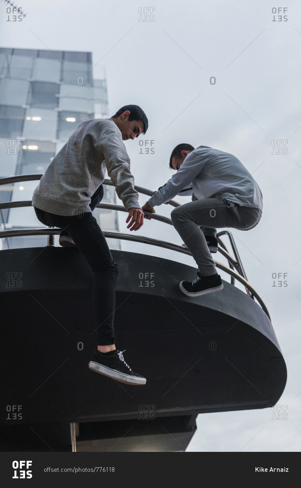 Two male friends in casual outfits hanging on railing of modern building while doing parkour in Madrid