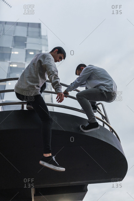 Two male friends in casual outfits hanging on railing of modern building while doing parkour in Madrid