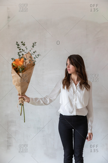 Lovely female in elegant outfit holding pretty flowers wrapped in paper while standing near shabby cement wall