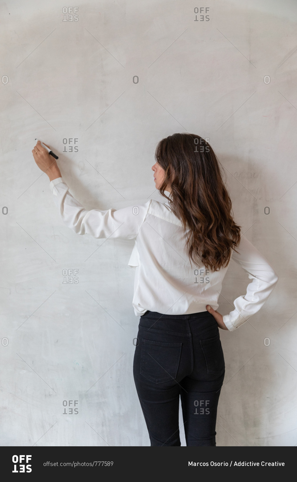 Back view of adult female in elegant outfit keeping hand on waist and writing on clean concrete wall with marker pen