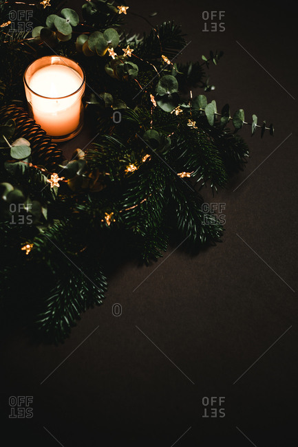 Small glass with flaming candle standing near green twigs and fresh cone of conifer tree on brown background