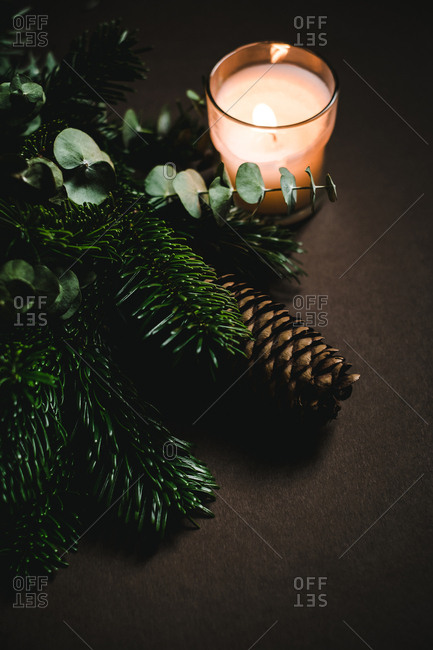 Small glass with flaming candle standing near green twigs and fresh cone of conifer tree on brown background