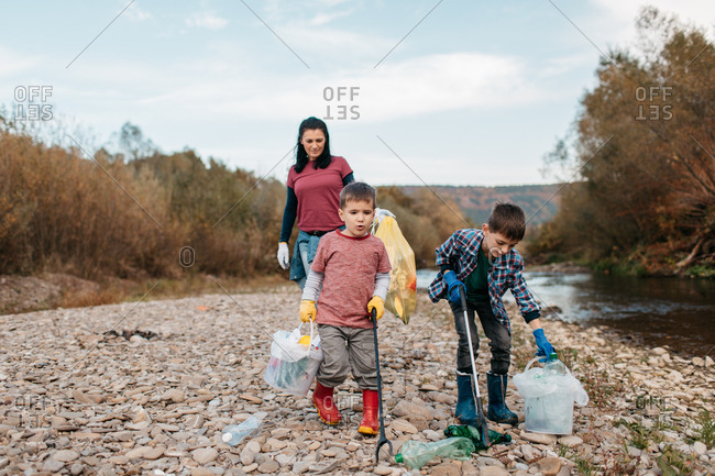 Two brothers with garbage pickers cleaning up plastic bottles in the nature. Children walking along river beach picking up plastic waste with their mother.