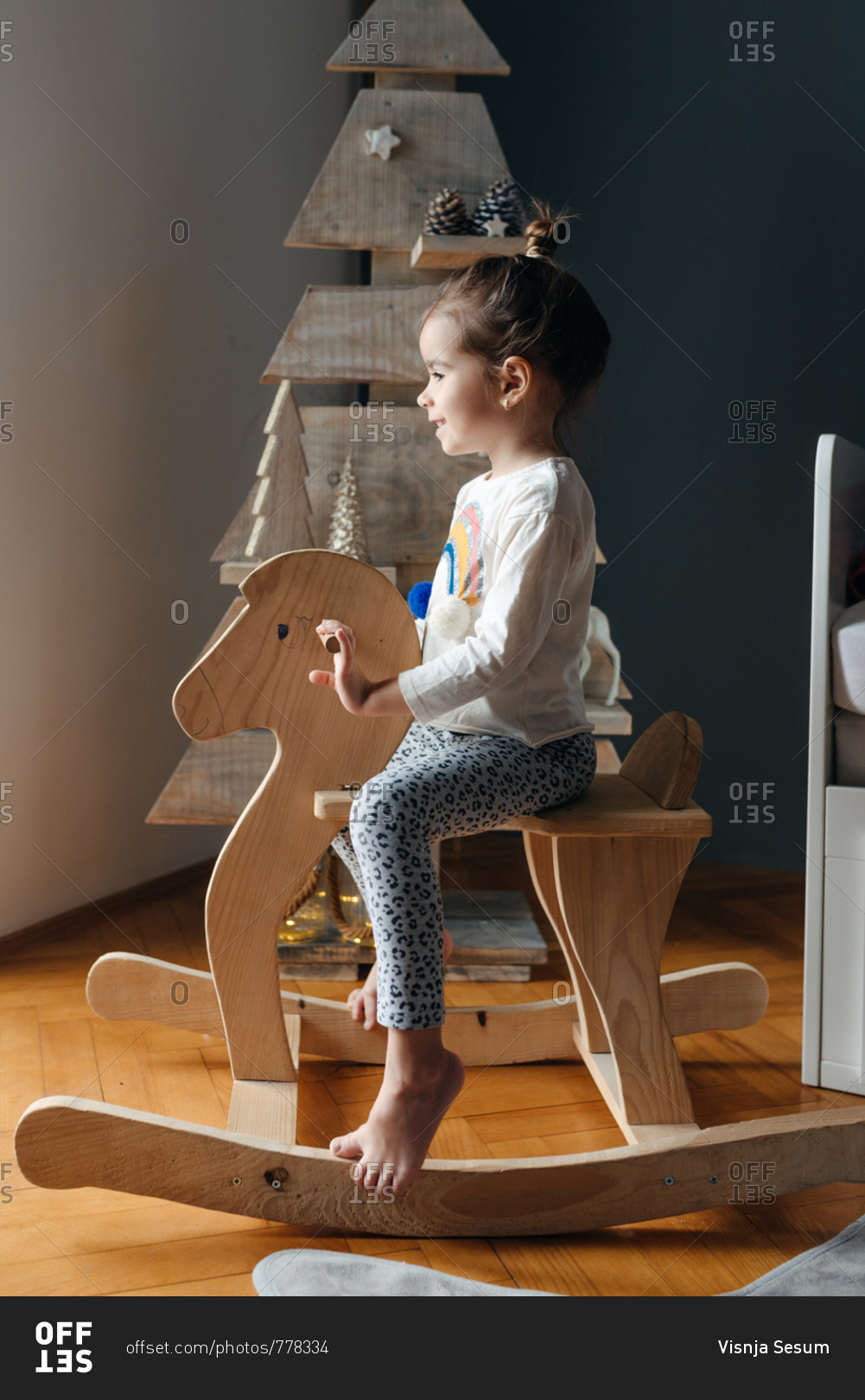 Side view of girl sitting on her wooden horse in front of a wooden Christmas tree