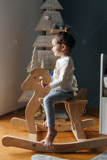 Side view of girl sitting on her wooden horse in front of a wooden Christmas tree