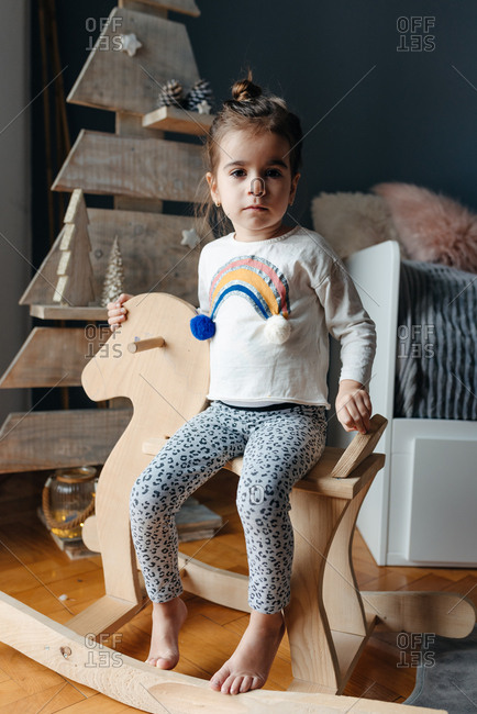 Little girl sitting on a wooden horse in front of a wooden Christmas tree