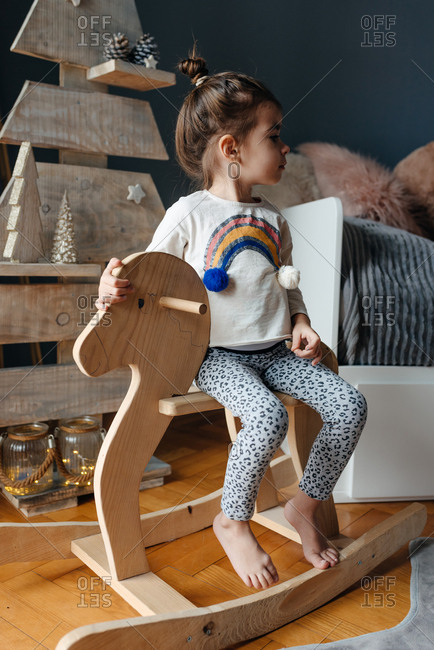 Little girl sitting on her wooden horse in front of a wooden Christmas tree