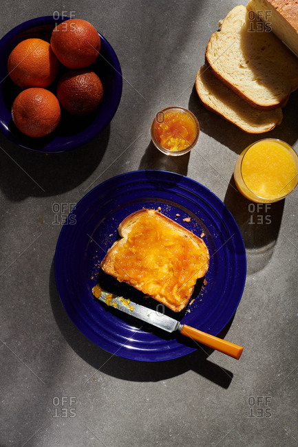 Orange Marmalade on toast on a blue plate with a butter knife on the plate beside blood oranges and orange juice