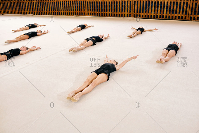 Group of little Caucasian female rhythmic gymnasts lying on mat and stretching while having workout at school gymnasium