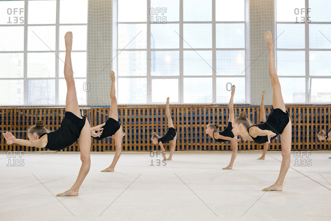 Group of little Caucasian female athletes standing in pose doing vertical splits while having rhythmic gymnastics class at school gymnasium