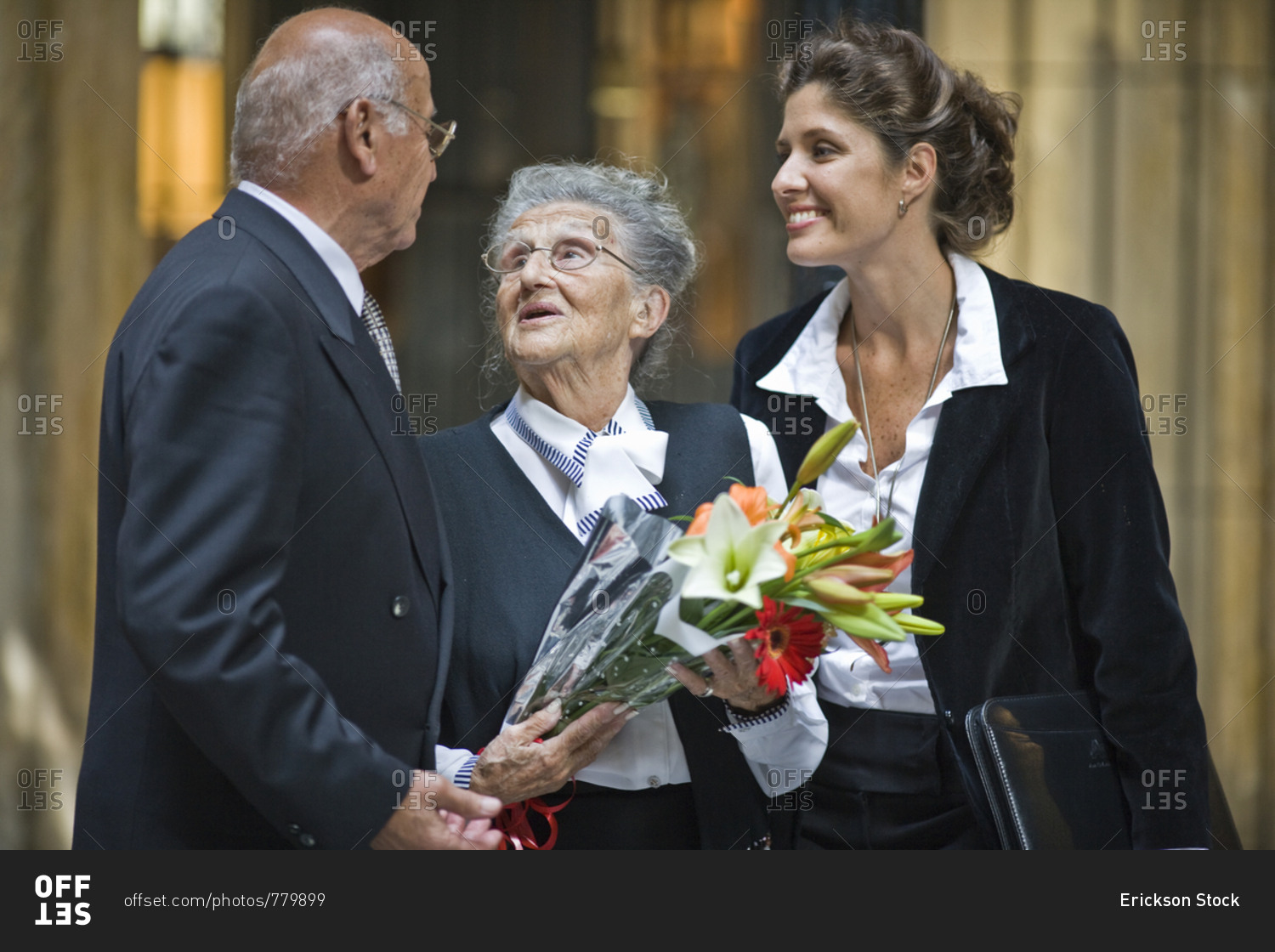 Senior woman holding flowers with her husband and mid-adult daughter while talking in a foyer.