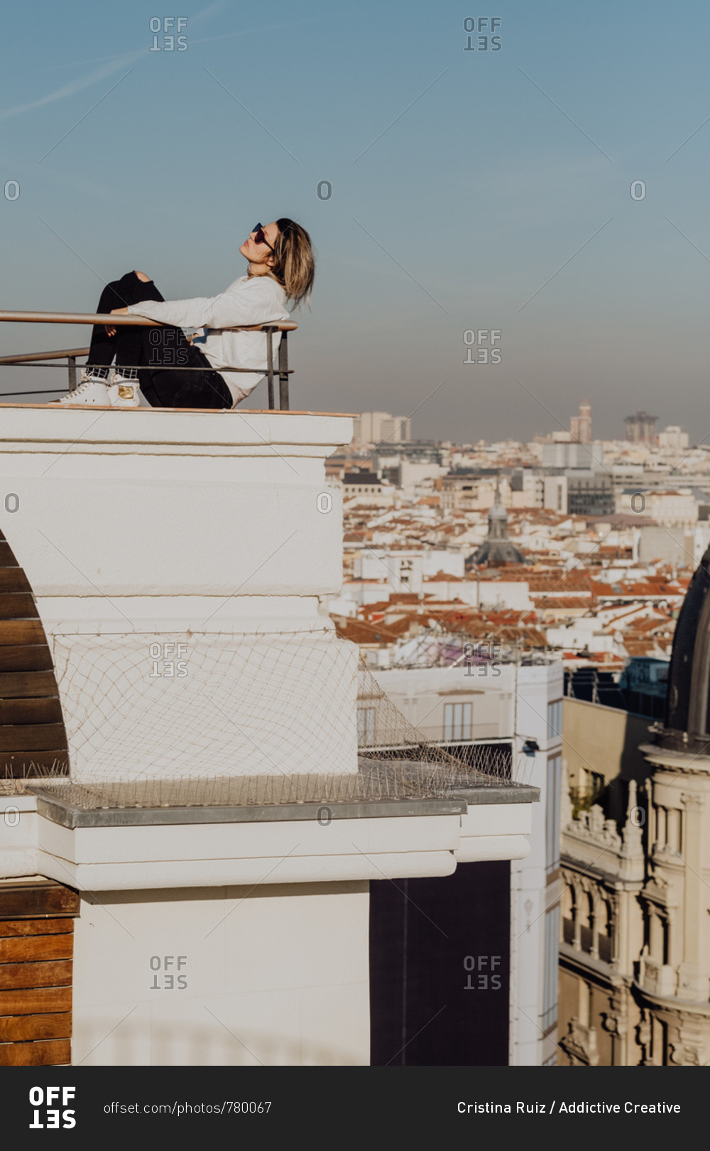 Side view of young stylish lady in casual wear and sunglasses leaning on rails on roof and blue sky on cityscape background in Madrid, Spain