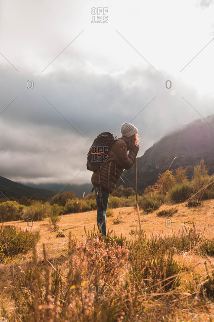 Side view of lady in hat and ski jacket with knapsack and walking stick walking on meadow near mountain in clouds in Isoba, Castile and Leon, Spain