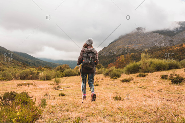 Back view of lady in hat and ski jacket with knapsack and walking stick walking on meadow near mountain in clouds in Isoba, Castile and Leon, Spain