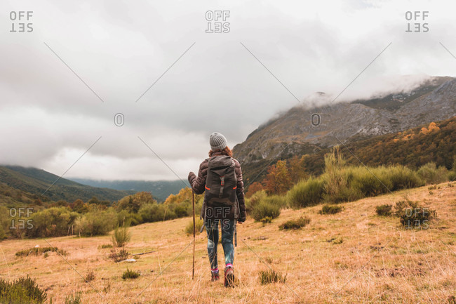 Back view of lady in hat and ski jacket with knapsack and walking stick walking on meadow near mountain in clouds in Isoba, Castile and Leon, Spain