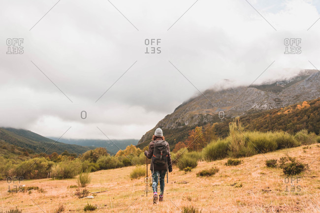 Back view of lady in hat and ski jacket with knapsack and walking stick walking on meadow near mountain in clouds in Isoba, Castile and Leon, Spain
