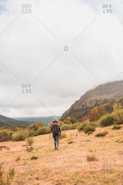 Back view of lady in hat and ski jacket with knapsack and walking stick walking on meadow near mountain in clouds in Isoba, Castile and Leon, Spain