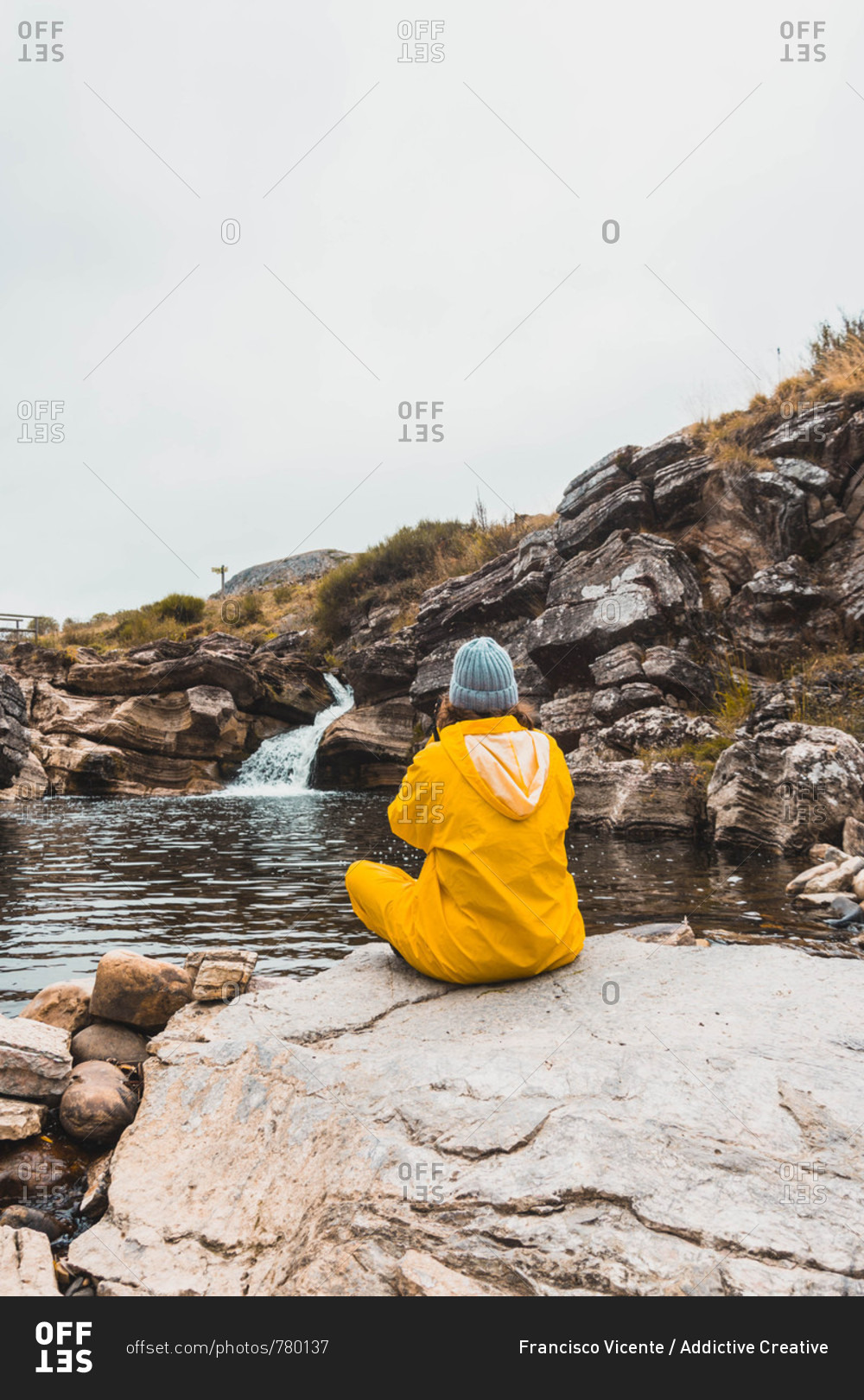 Back view of woman in hat and yellow raincoat sitting in stones and shooting rift of river between rock hills on camera in Isoba, Castile and Leon, Spain