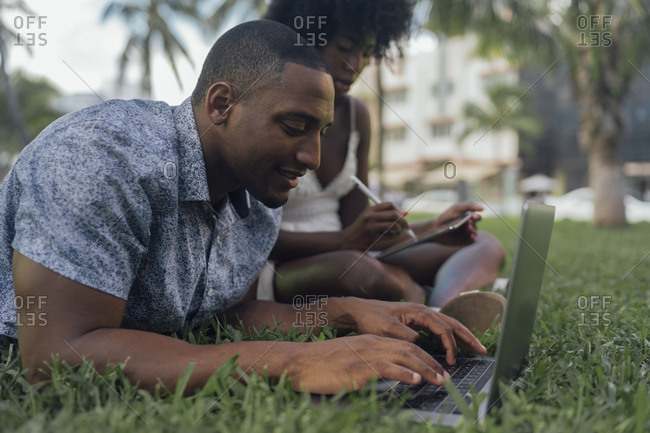 USA- Florida- Miami Beach- young couple using tablet and laptop on lawn in a park