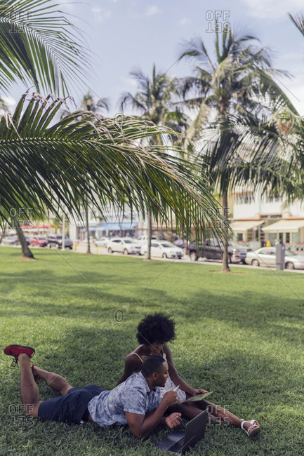 USA- Florida- Miami Beach- young couple using tablet and laptop on lawn in a park