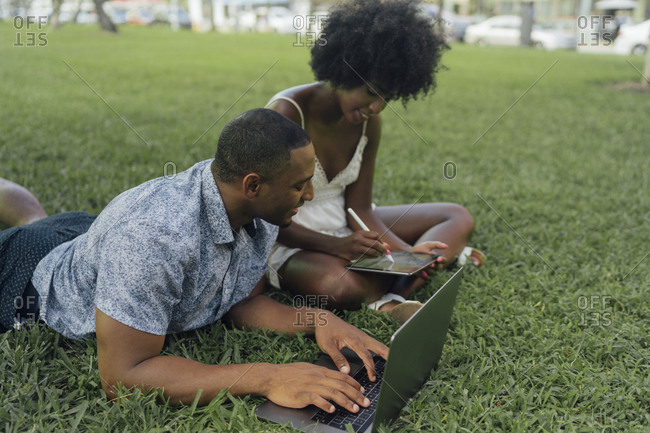 Young couple using tablet and laptop on lawn in a park