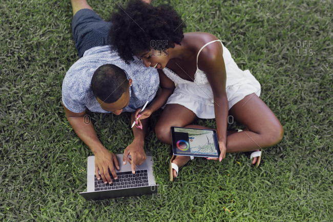 Young couple using tablet and laptop on lawn in a park