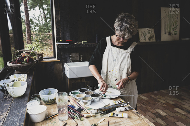 Senior woman wearing glasses, black top and white apron standing in studio, mixing colours.