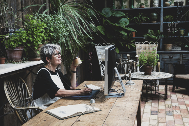 Senior woman wearing glasses, black top and white apron sitting at a wooden table, working on desktop computer.