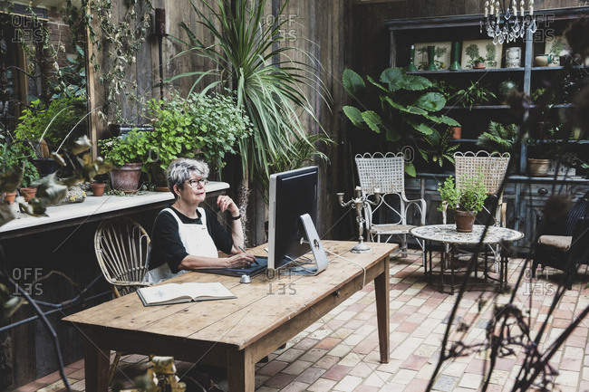 Senior woman wearing glasses, black top and white apron sitting at a wooden table, working on desktop computer.