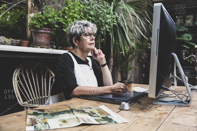 Senior woman wearing glasses, black top and white apron sitting at a wooden table, working on desktop computer.